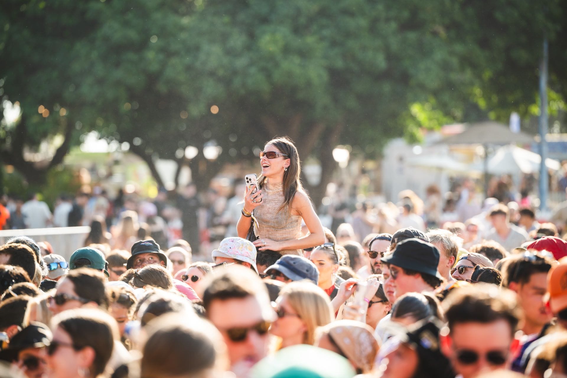 A woman stands above a large crowd at an outdoor event, holding a phone, with trees in the background.