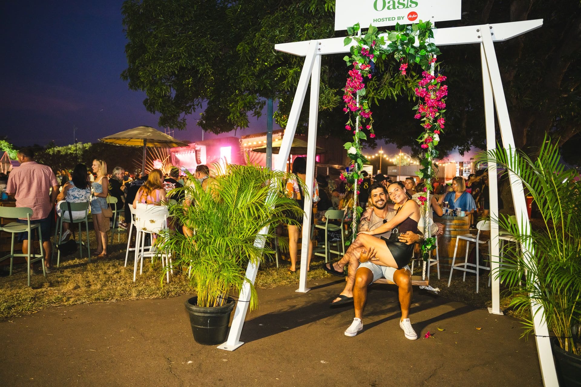 A couple sits on a decorated swing at an outdoor event, surrounded by people, plants, and lights in the evening.
