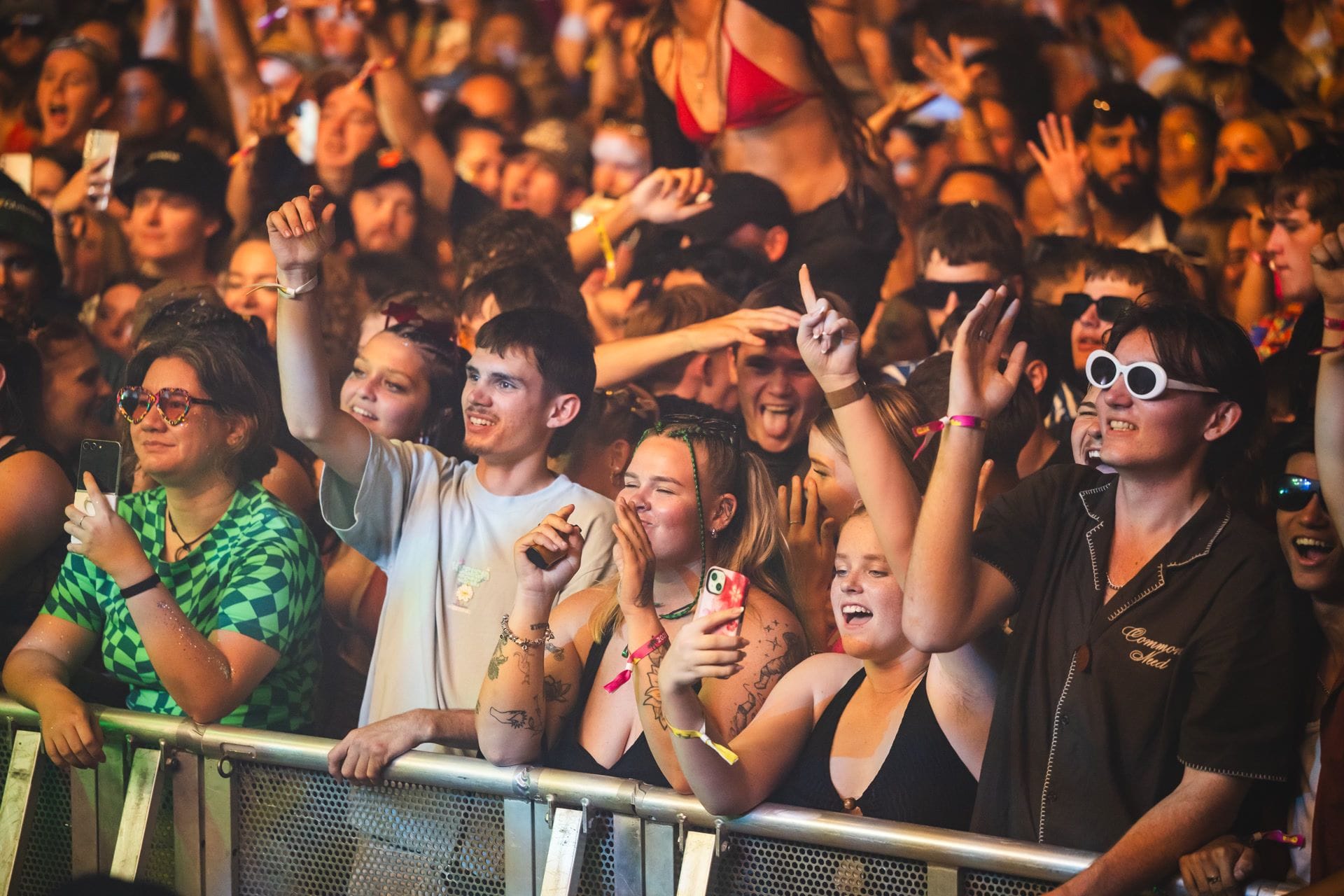 A lively crowd at a concert, with people smiling, cheering, and holding phones. Some wear sunglasses and colorful outfits, enjoying the event.