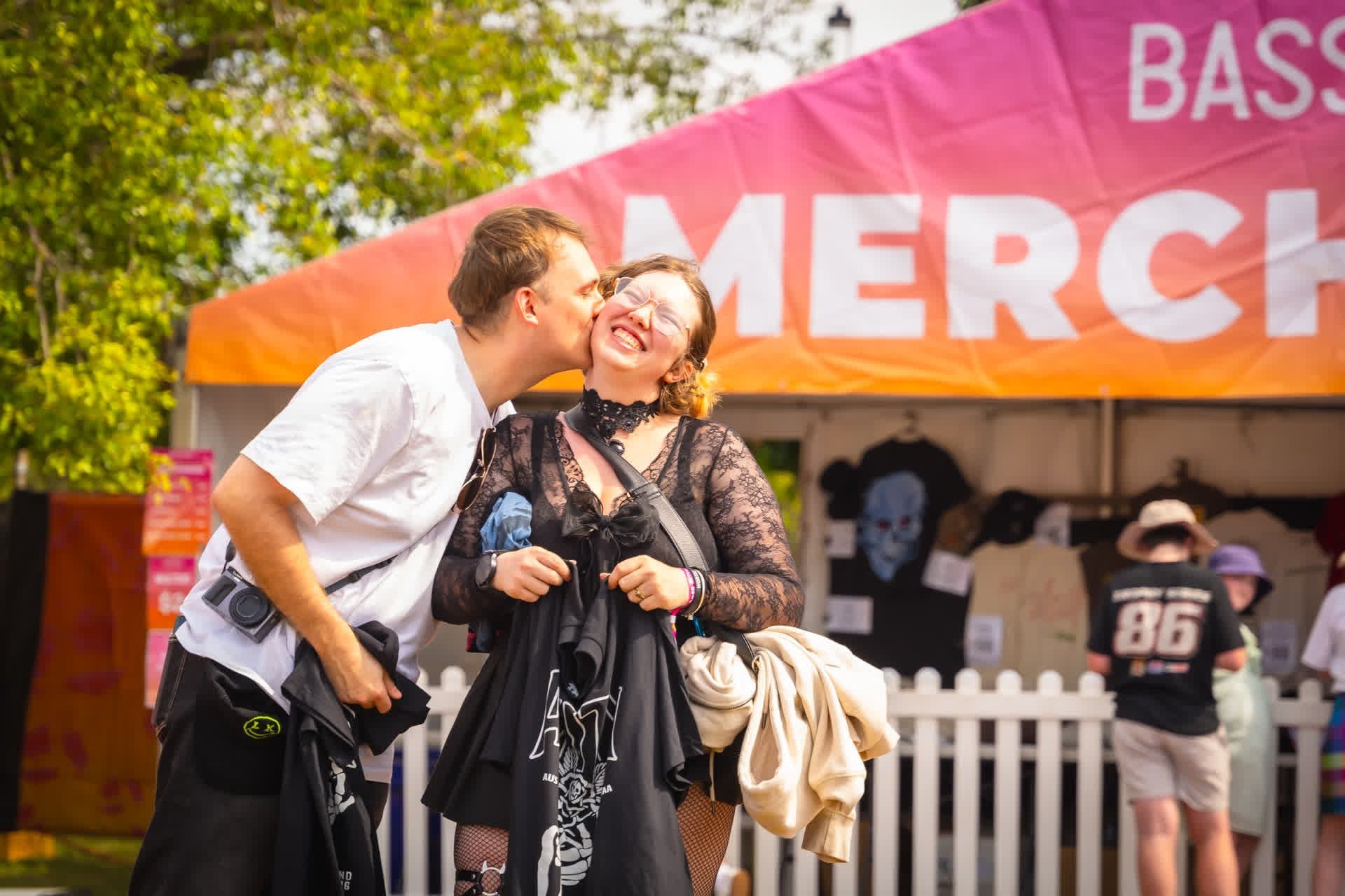 A person playfully kisses another on the cheek outside a merchandise tent at a festival. They hold T-shirts and smile, surrounded by a lively crowd.