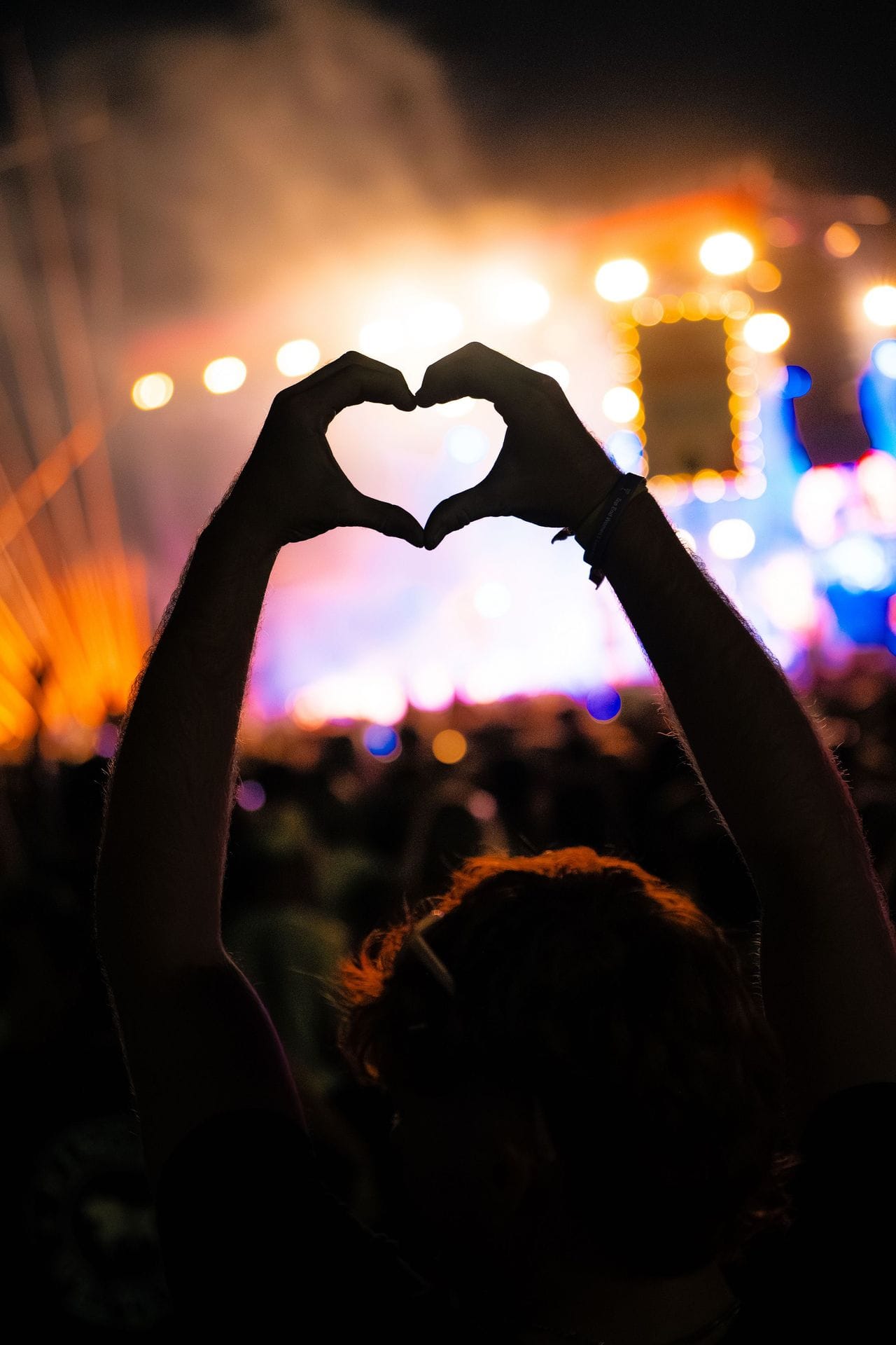 Person forming a heart shape with hands at a concert, bright stage lights and blurred crowd in the background.
