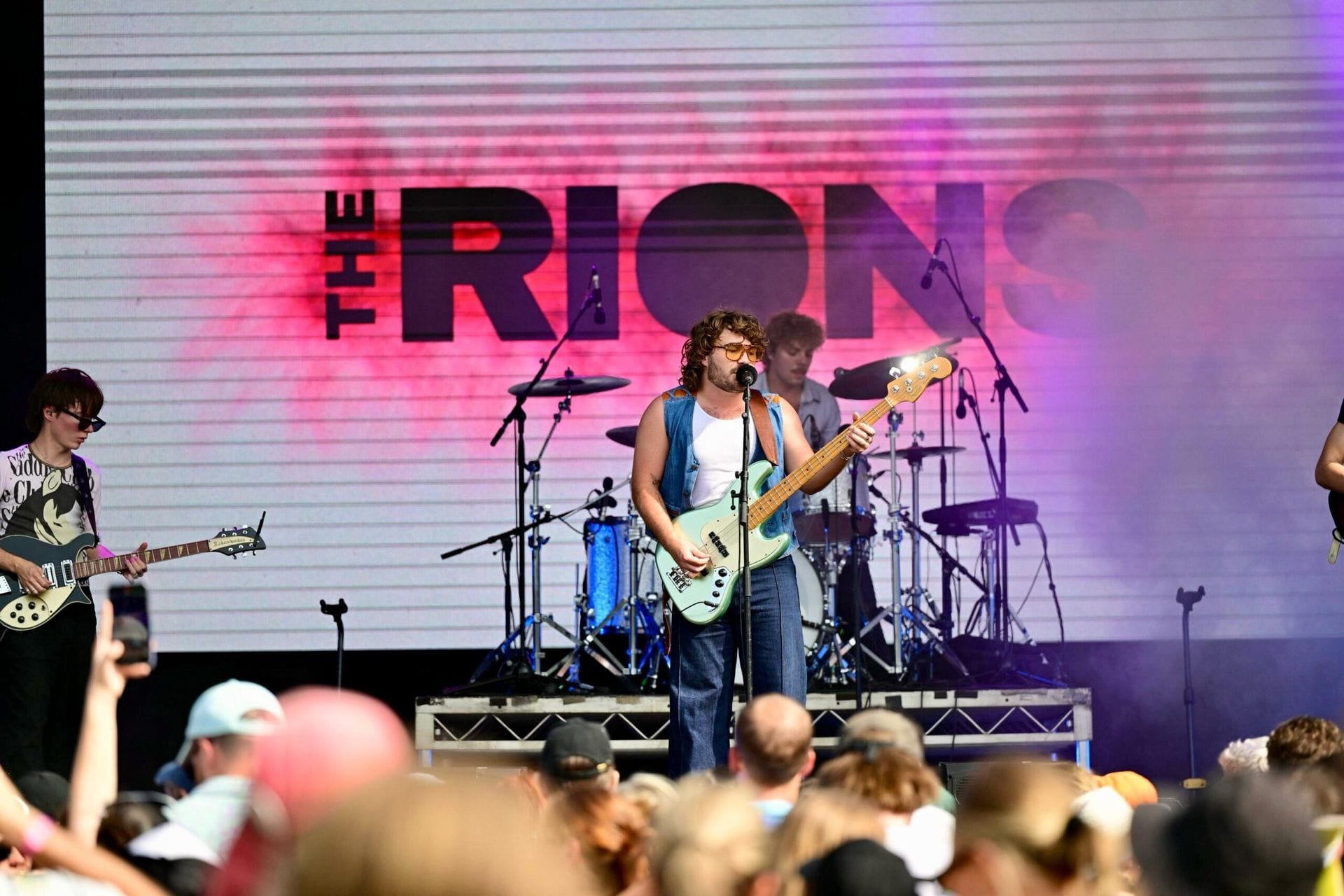 A band performs energetically on stage with guitars and drums, in front of a crowd. The backdrop displays "The Rions" in bold letters.
