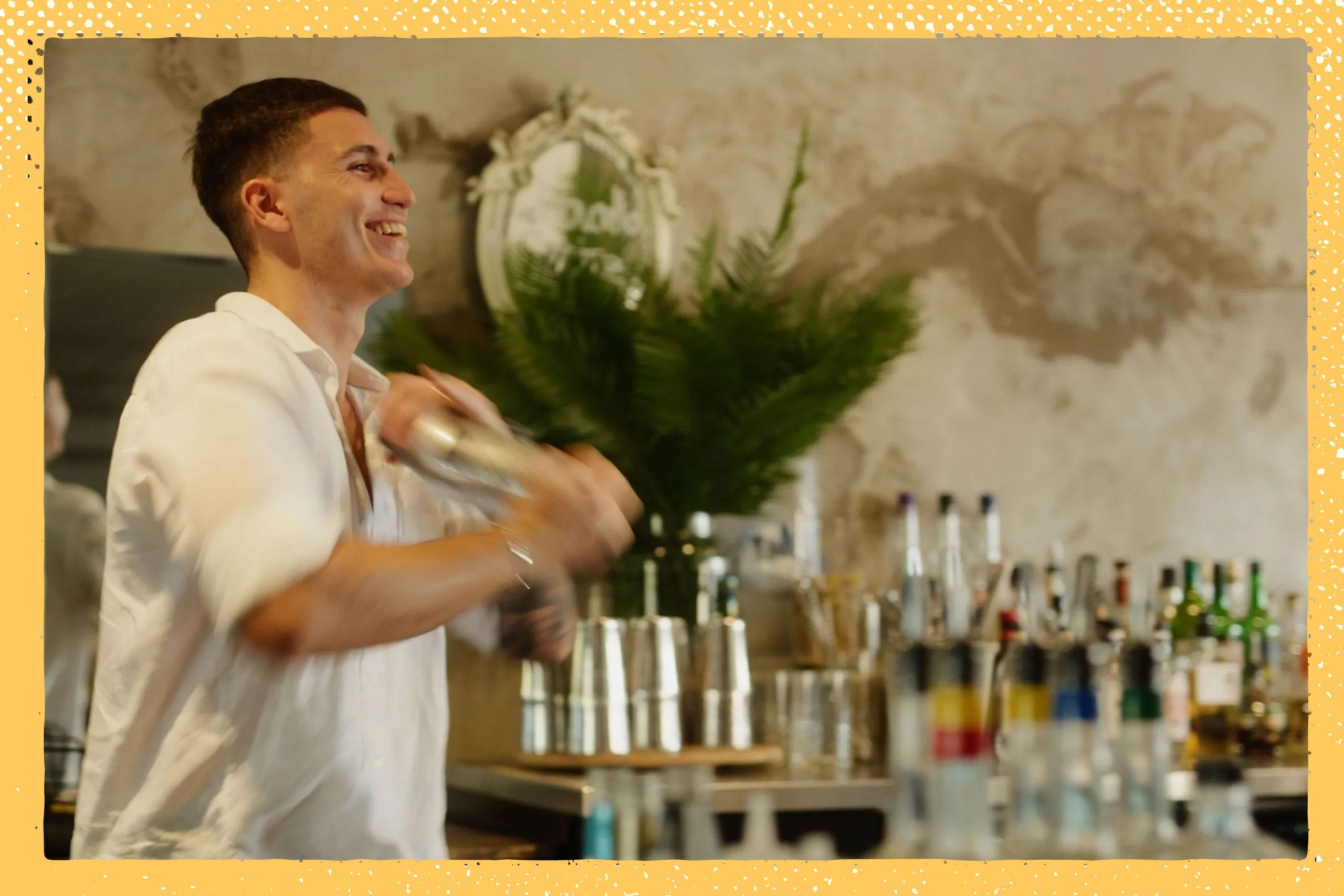 Bartender in a white shirt smiling while shaking a cocktail shaker, with bottles and greenery in the background.