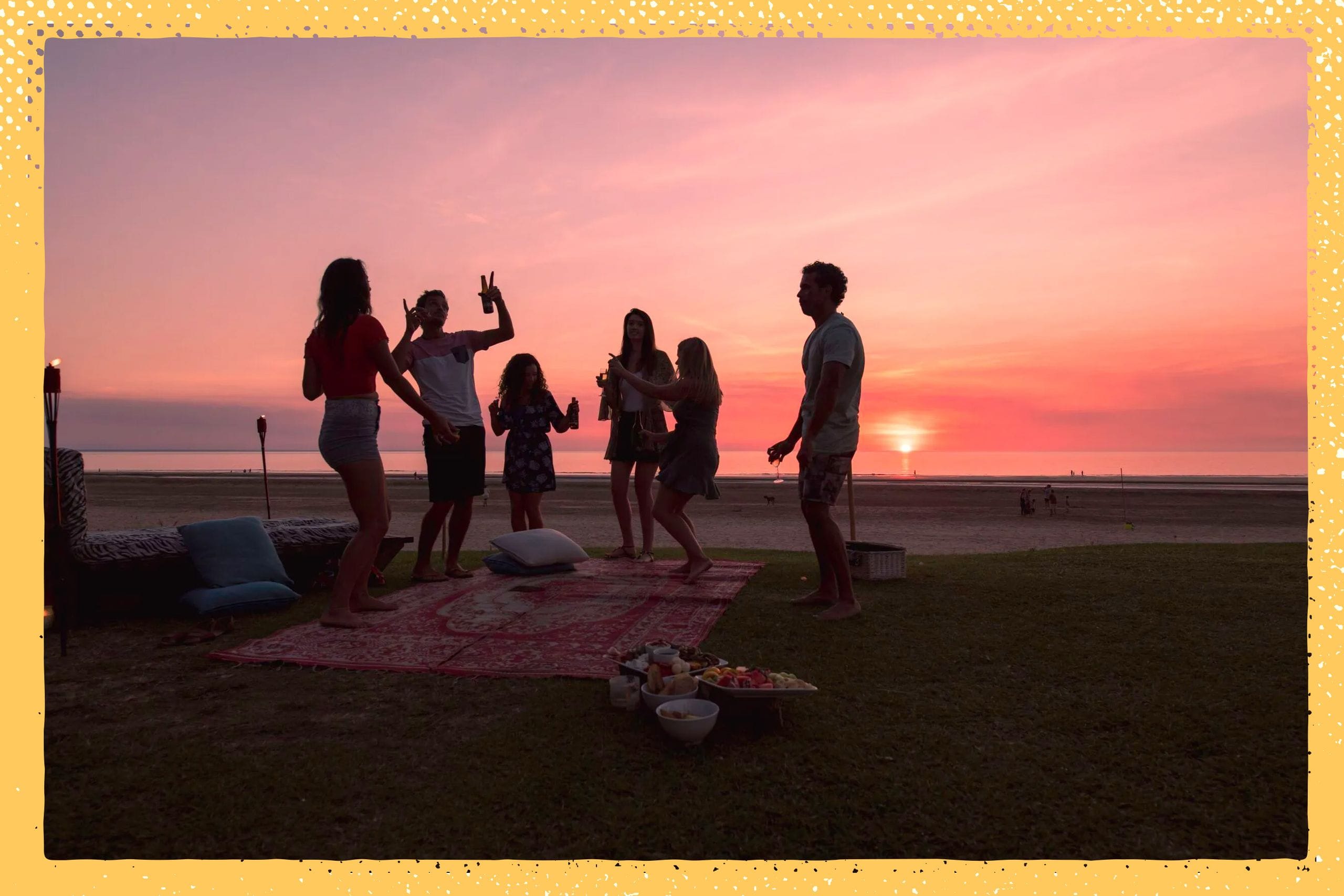 Silhouetted group dancing on a beach at sunset, with a picnic setup on a blanket and a vibrant pink sky.