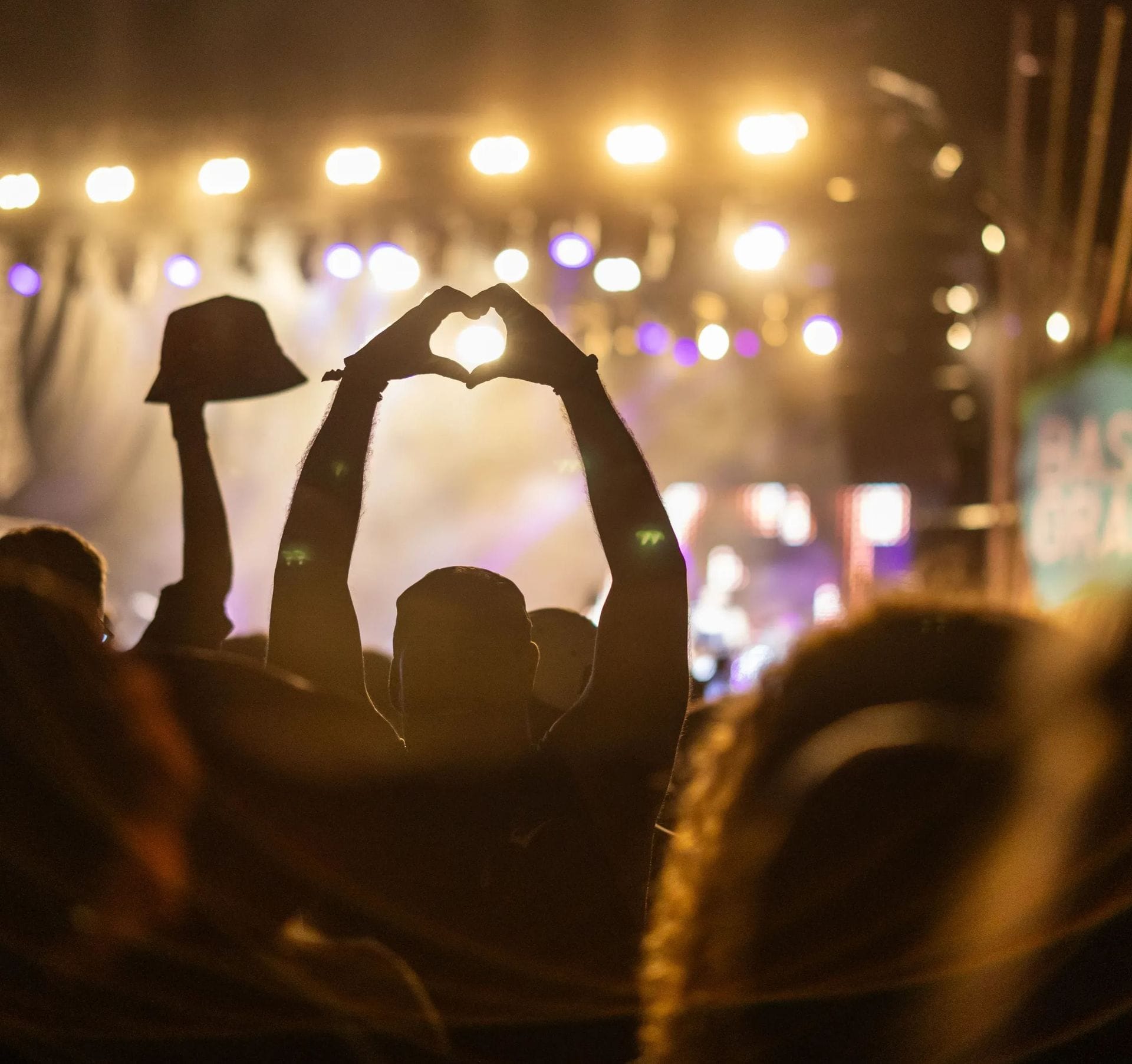 Silhouetted crowd at a concert, with one person forming a heart shape with hands against bright stage lights.