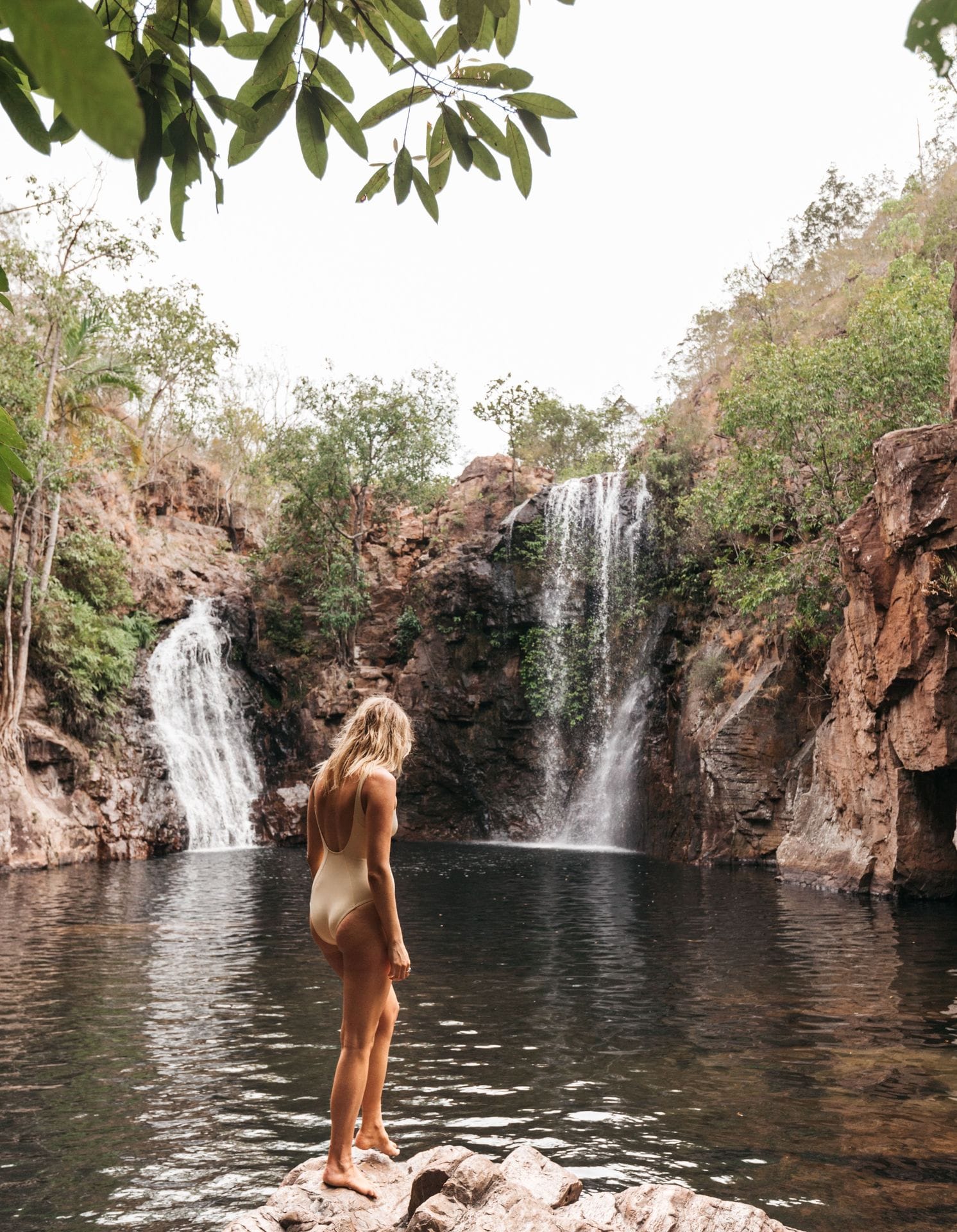 A person in a swimsuit stands on a rock, overlooking a tranquil pool with two cascading waterfalls surrounded by lush greenery.