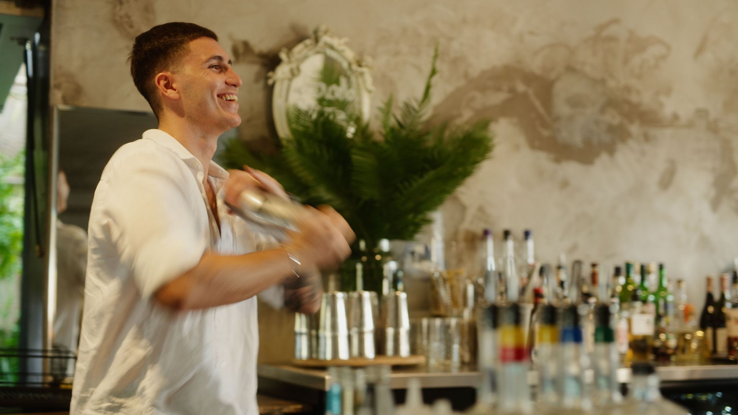Bartender in a white shirt shaking a cocktail shaker, standing behind a bar with various bottles and a plant in the background.