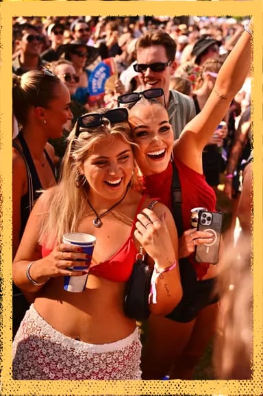 Two women smiling and posing at an outdoor festival, surrounded by a crowd. One holds a drink, the other raises her arm, both wearing sunglasses.