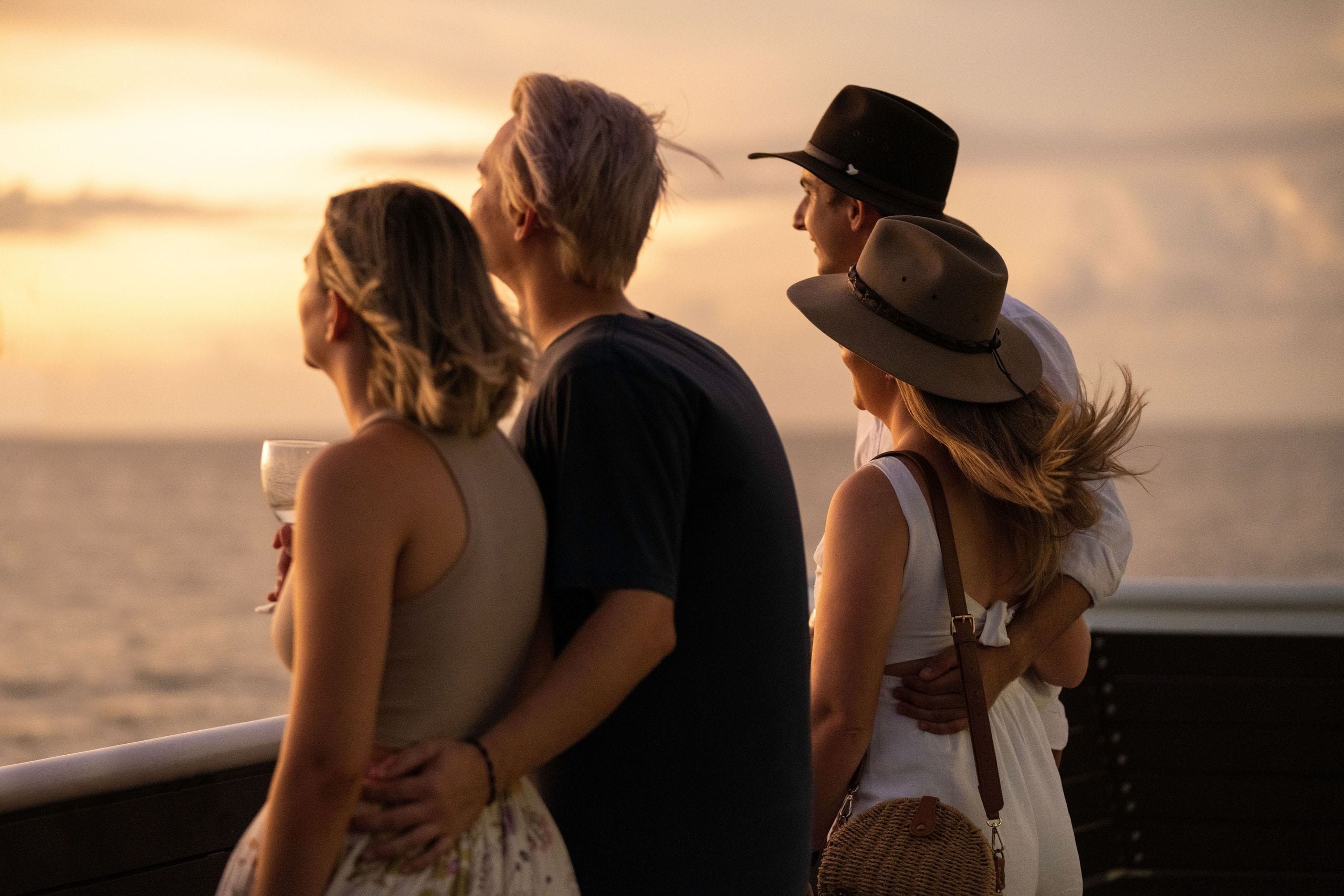 Four people on a deck at sunset, two wearing hats, embracing and enjoying the ocean view. One holds a wine glass.