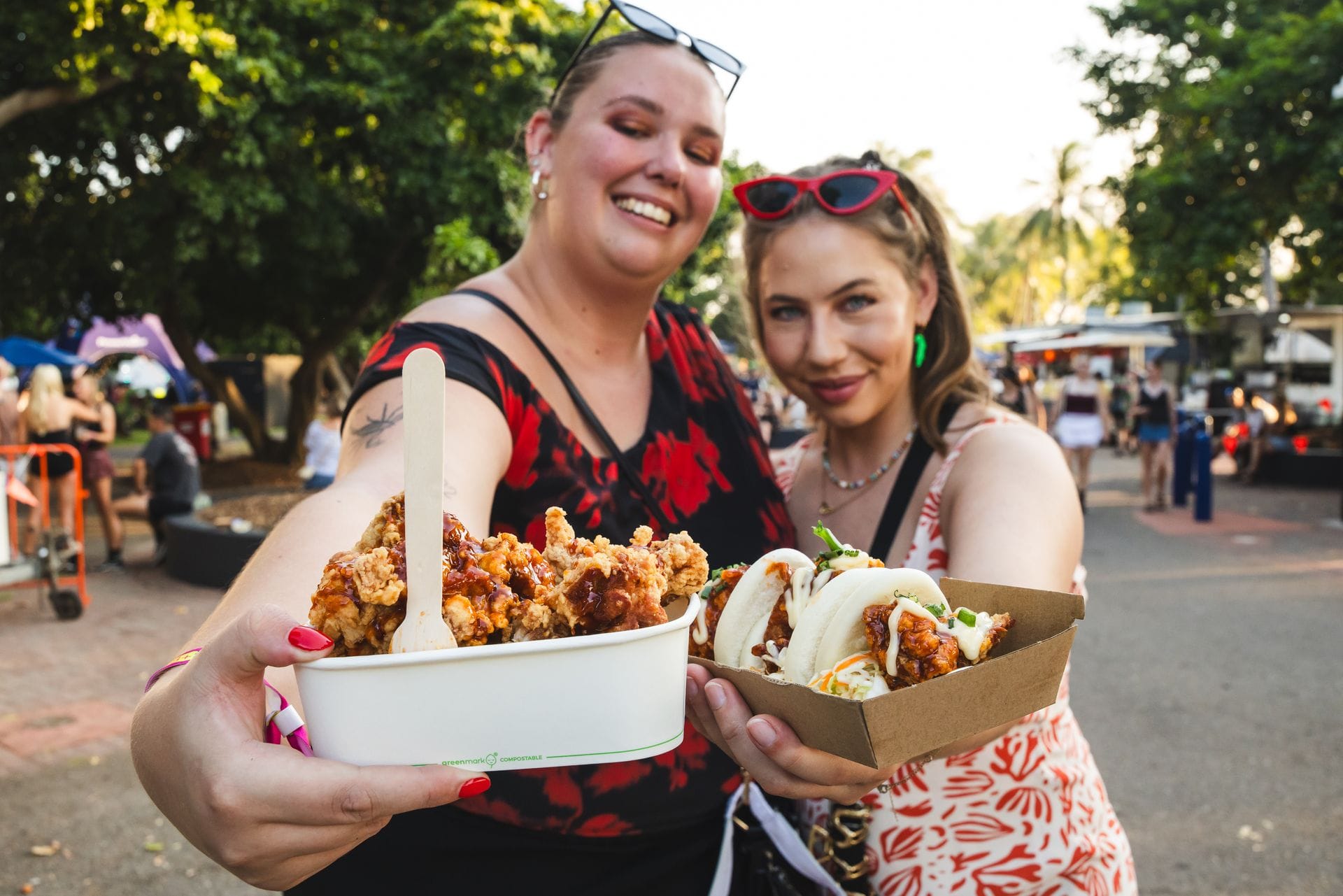 Two smiling women holding boxes of fried chicken and tacos at an outdoor food market, with trees and people in the background.