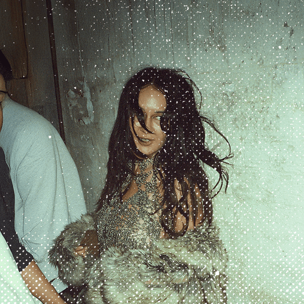 Woman with long hair in a textured dress and fur coat stands against a worn wall, smiling slightly.