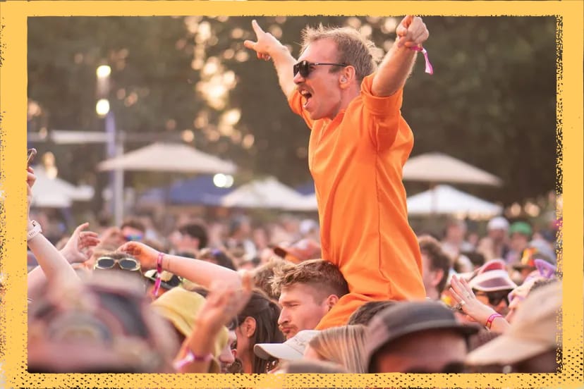 Man in orange shirt cheering on shoulders in a lively outdoor festival crowd, with trees and umbrellas in the background.