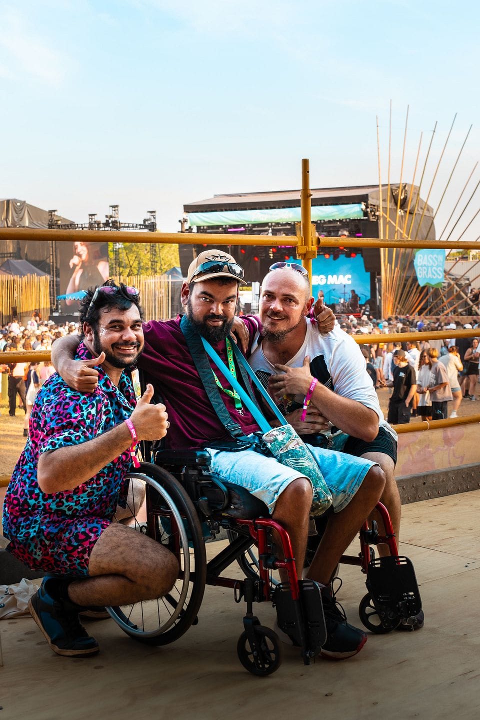 Three men smiling at an outdoor music festival, with one in a wheelchair. They are giving thumbs up, with a stage and crowd in the background.