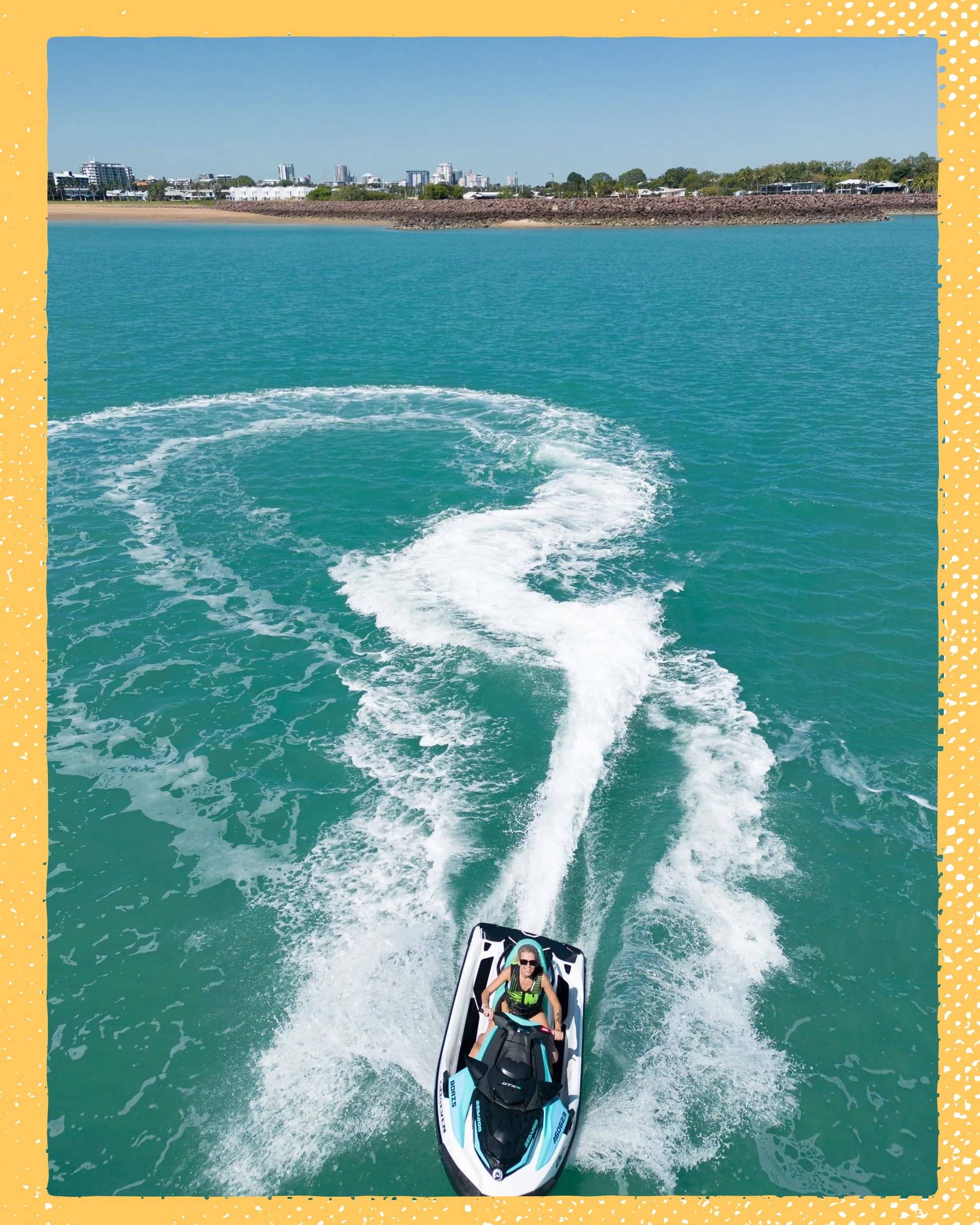 Person riding a jet ski on turquoise water, creating a circular wake pattern. Coastline and cityscape visible in the background.