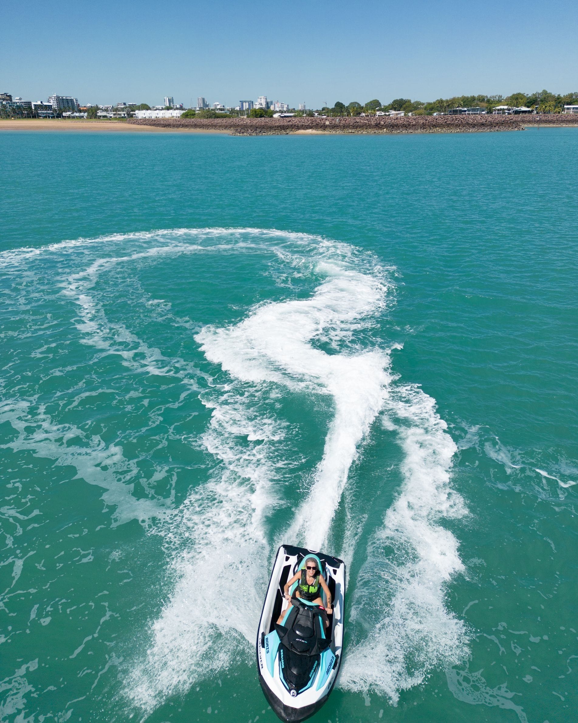 Person riding a jet ski on clear turquoise water, creating a circular wake pattern, with a distant shoreline under a clear blue sky.