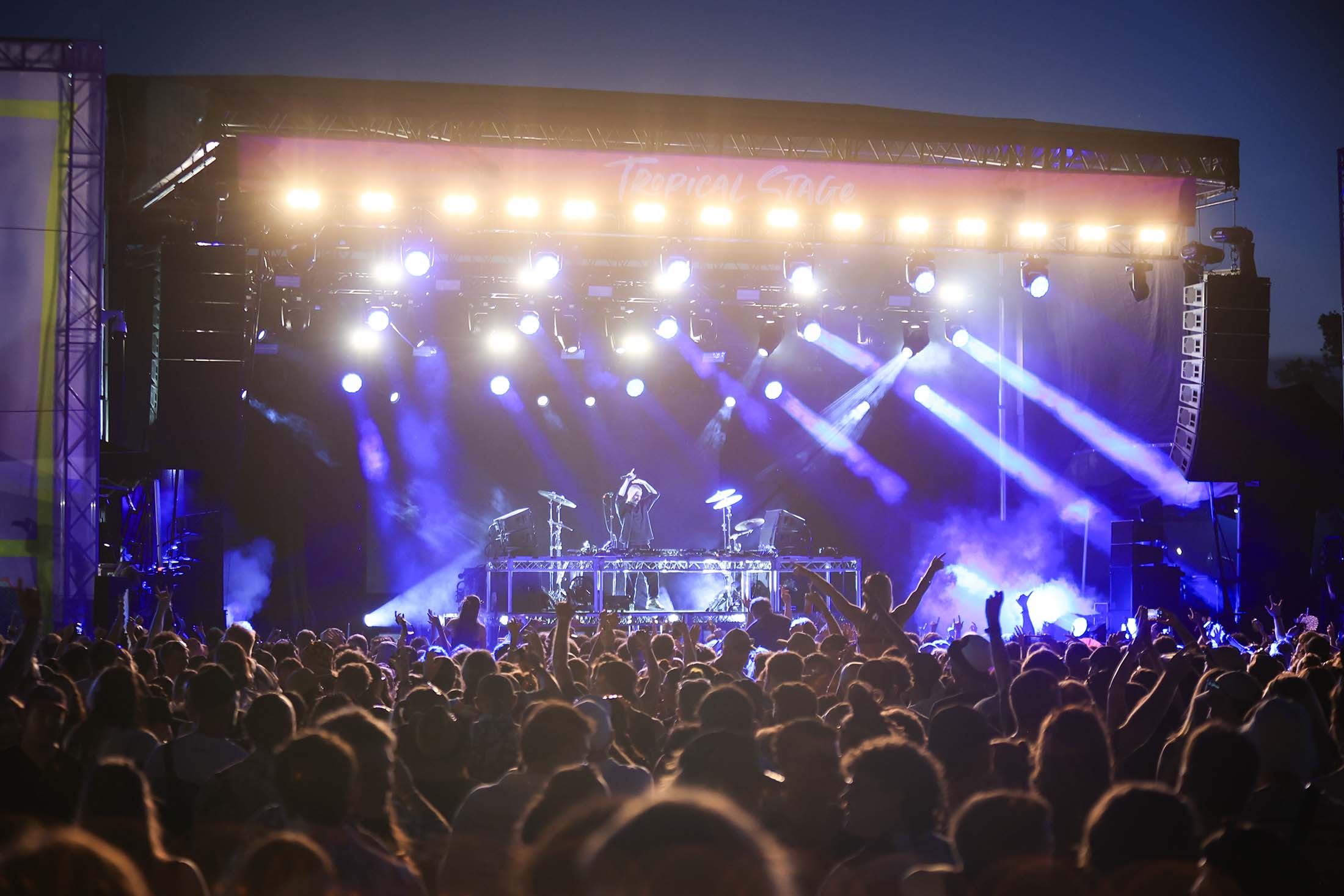 DJ performs on 'Tropical STAGE' at a night concert, stage lit with bright blue beams and smoke, a large crowd with hands up.