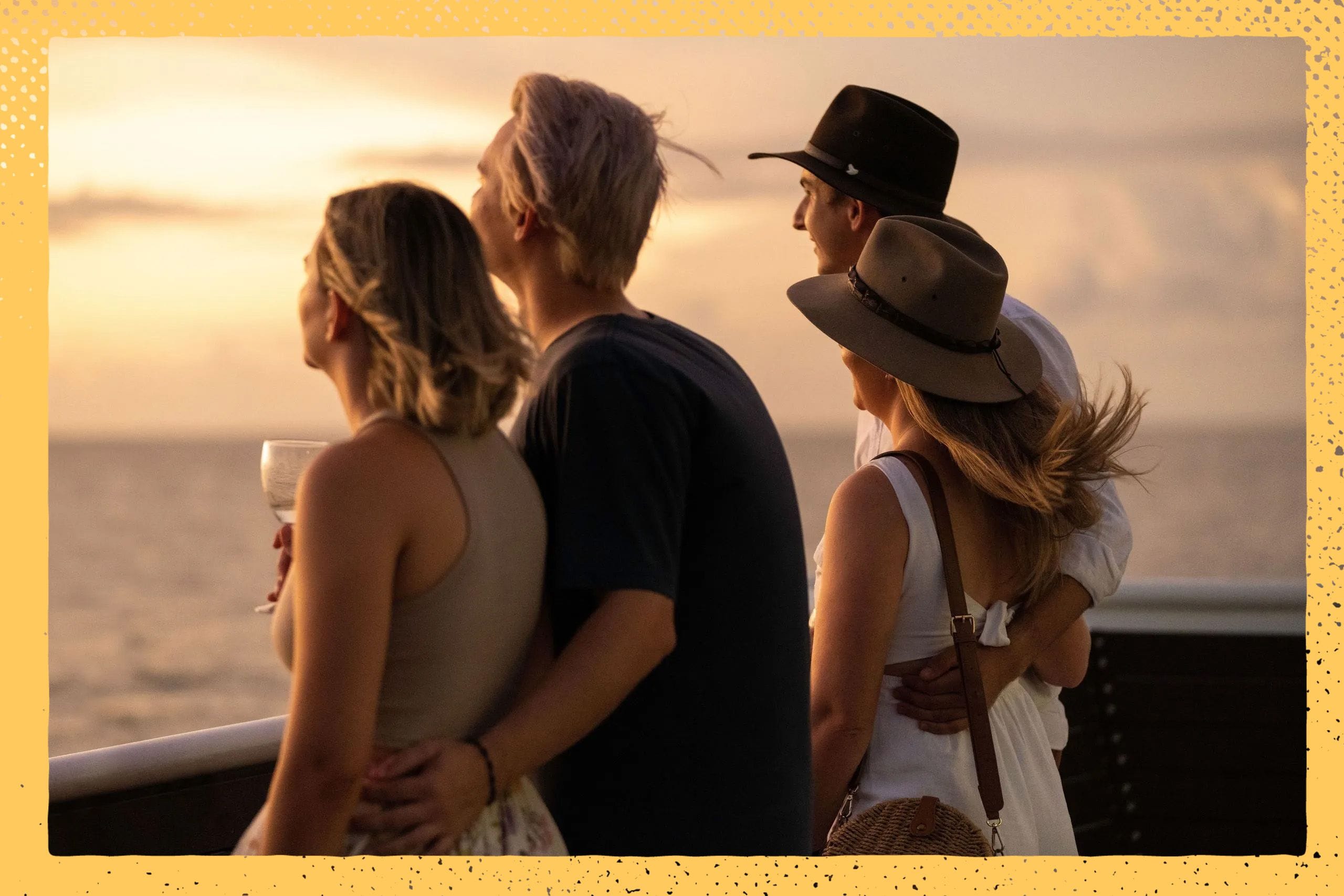 Two couples stand on a deck, gazing at the ocean during sunset. They are wearing casual summer clothes and hats, with one holding a drink.