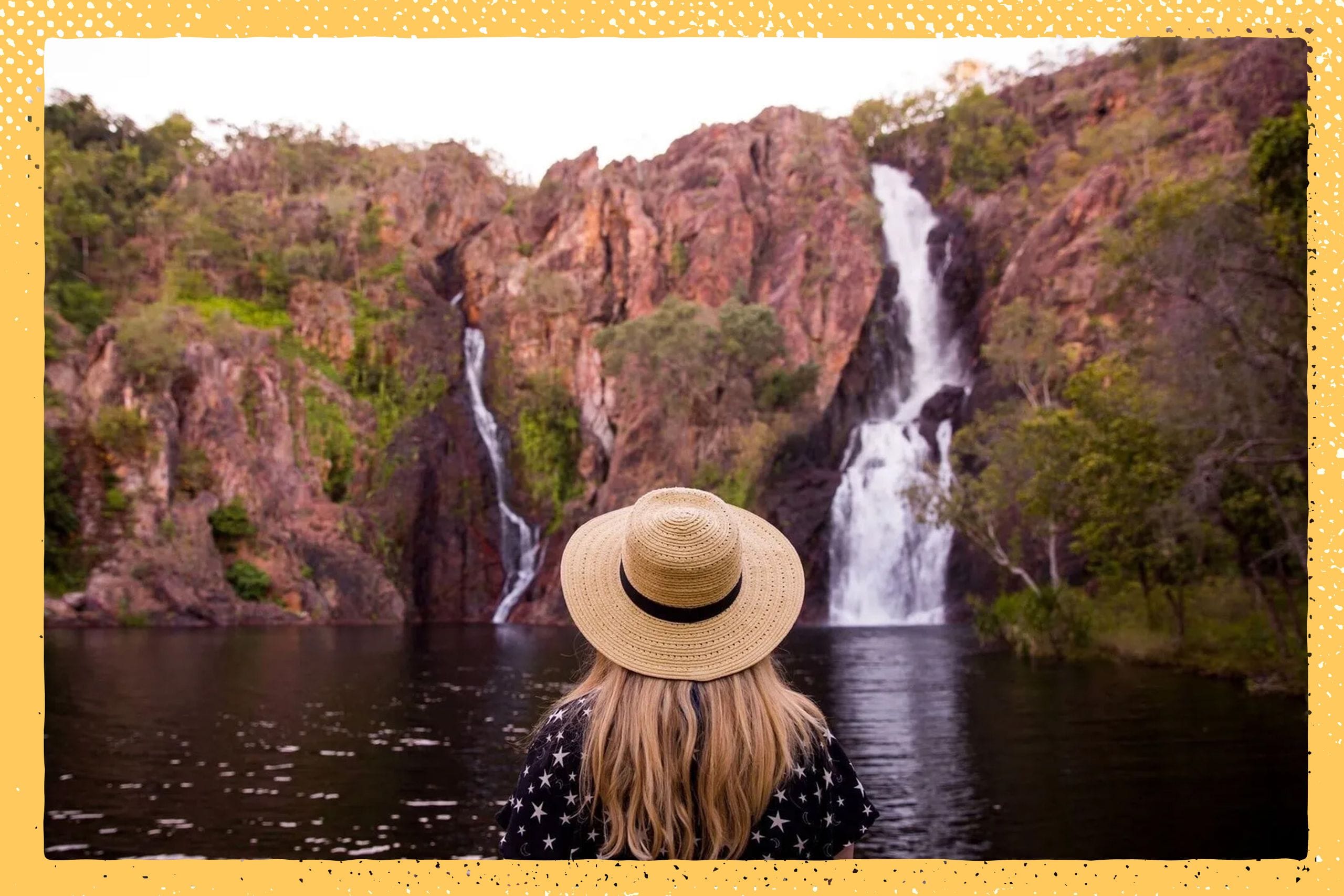 Person in a straw hat stands by a lake, facing a scenic waterfall cascading down a rocky cliff surrounded by greenery.