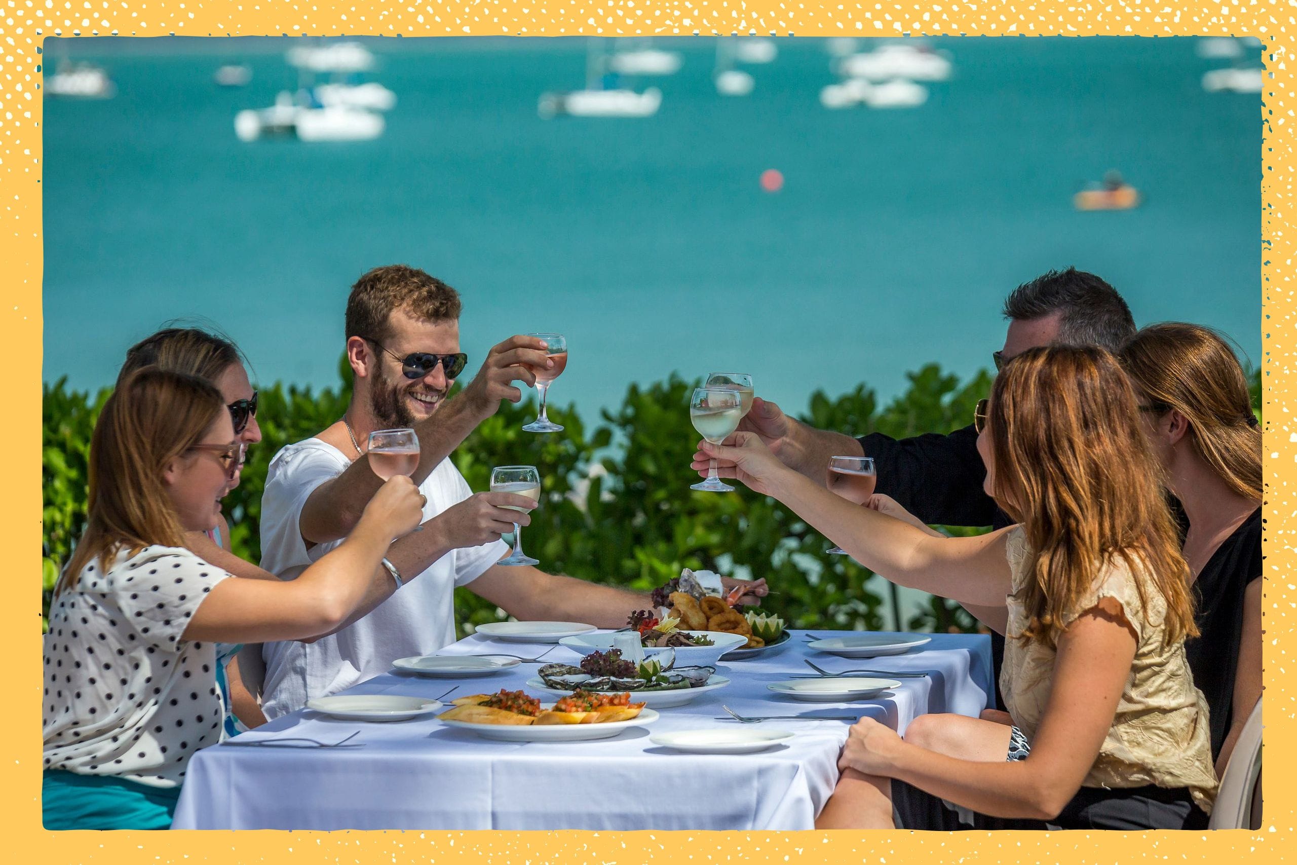 Group of people toasting with wine glasses at an outdoor dining table by the sea, with boats visible in the background.