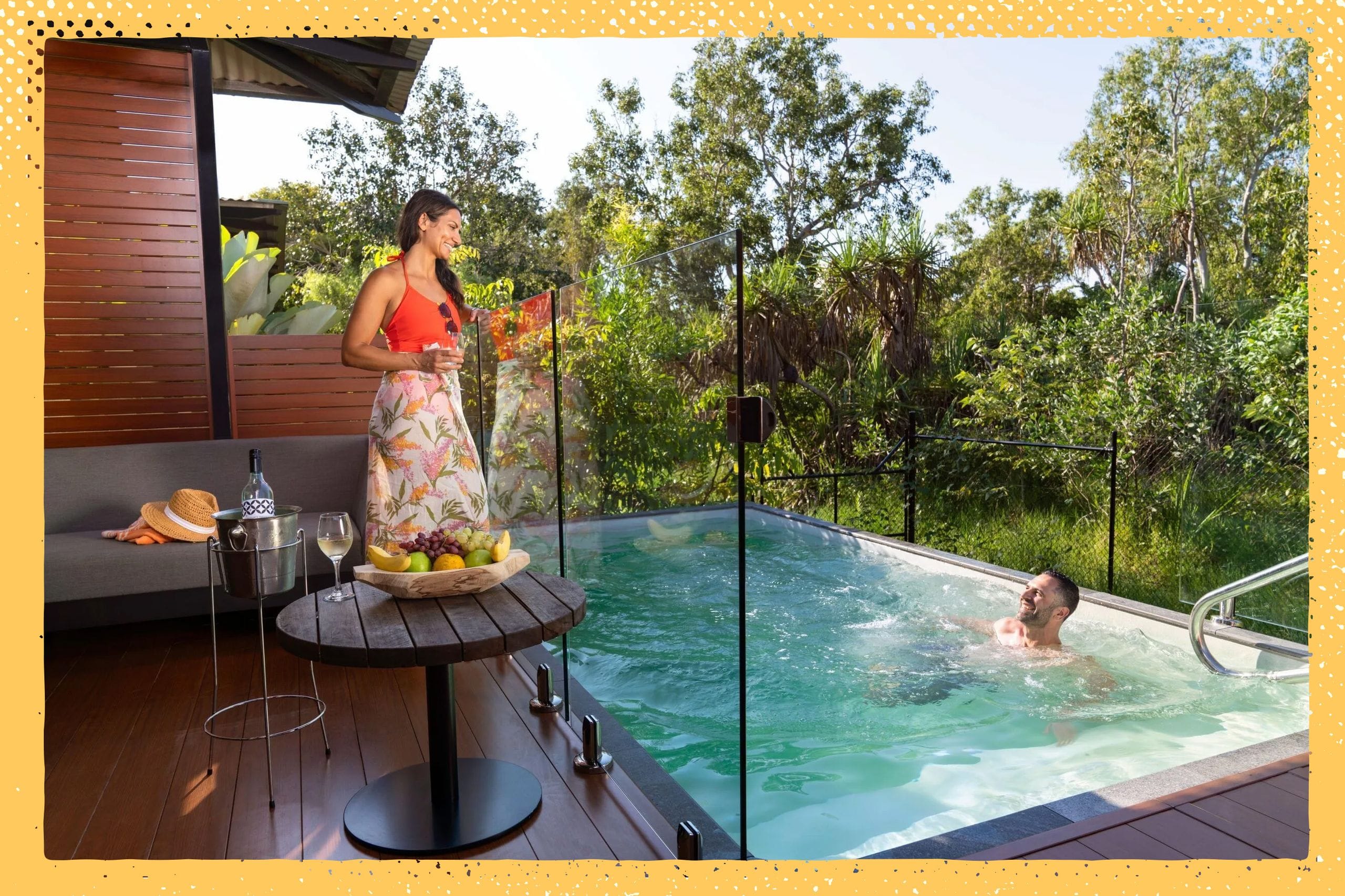 Woman in a red top stands by a poolside table with fruit and wine, watching a man swim. Surrounded by greenery and wooden decking.