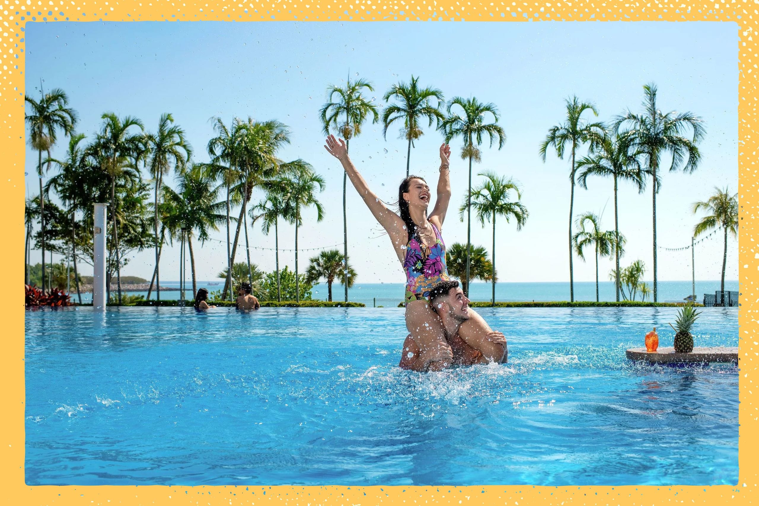 A woman joyfully raises her arms while sitting on a man's shoulders in a pool, surrounded by palm trees and a view of the ocean.
