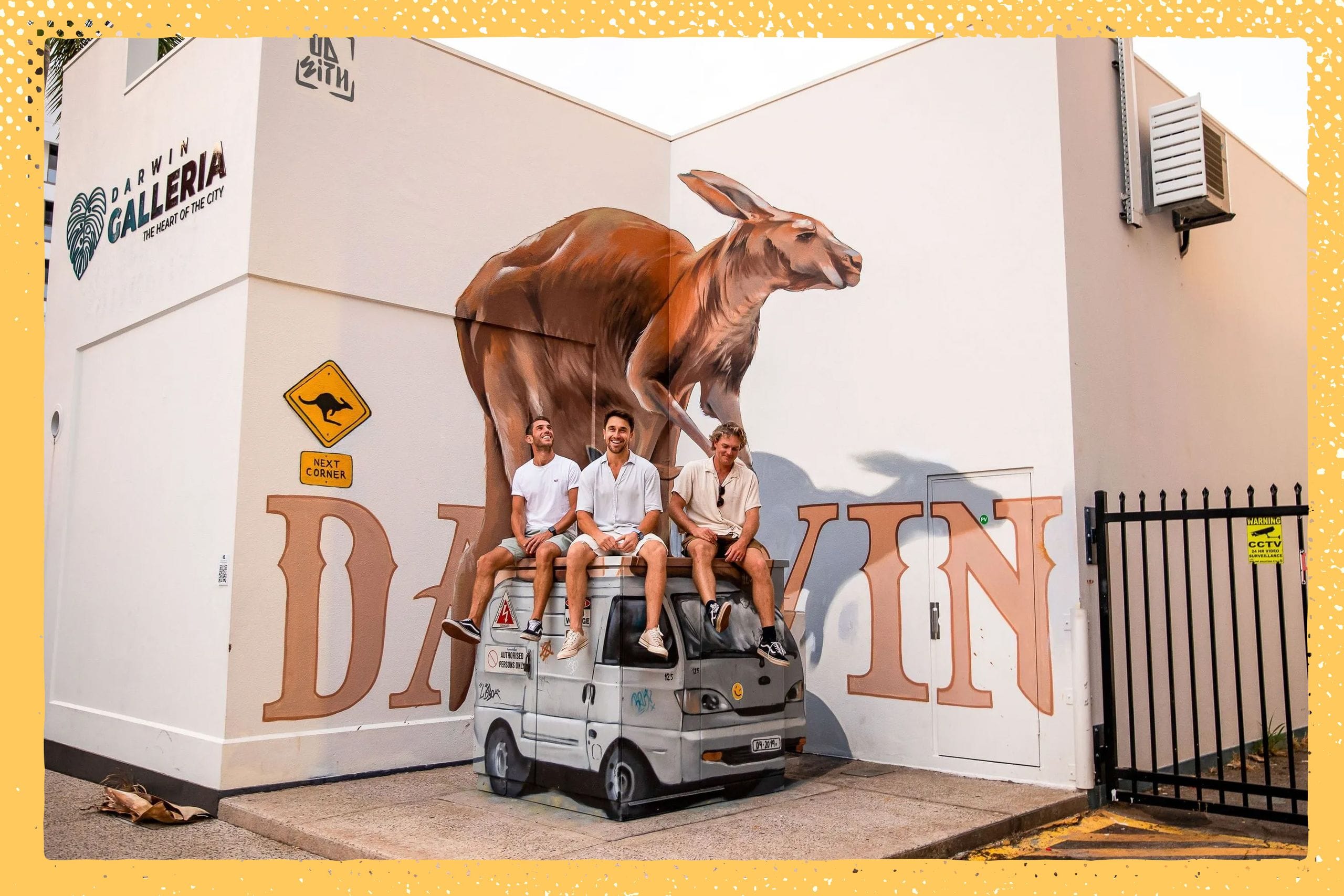 Three people sit on a utility vehicle in front of a large kangaroo mural on a building wall, with "DARWIN" painted beneath.