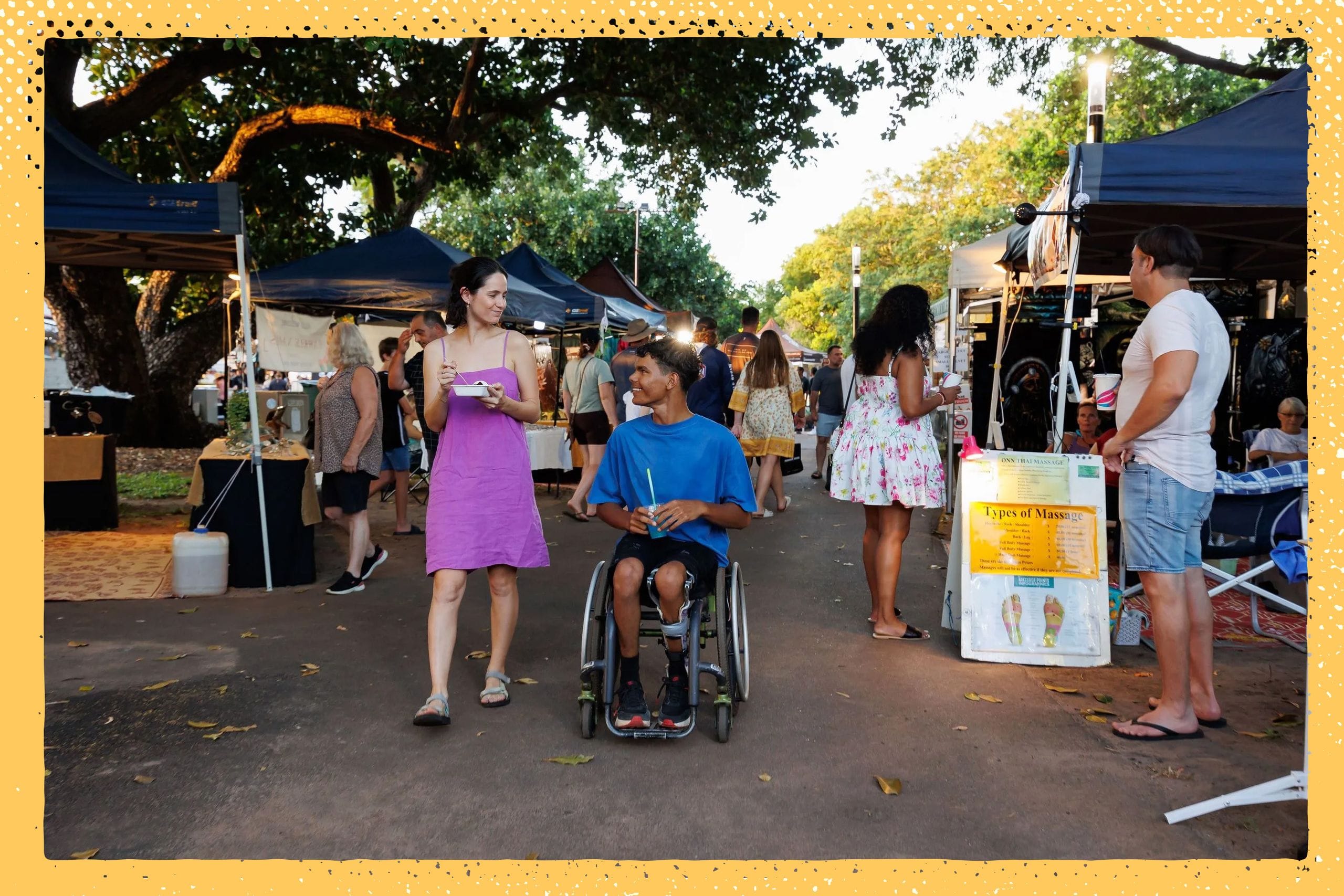 A woman and a man in a wheelchair enjoy a lively outdoor market with various stalls and people in the background.