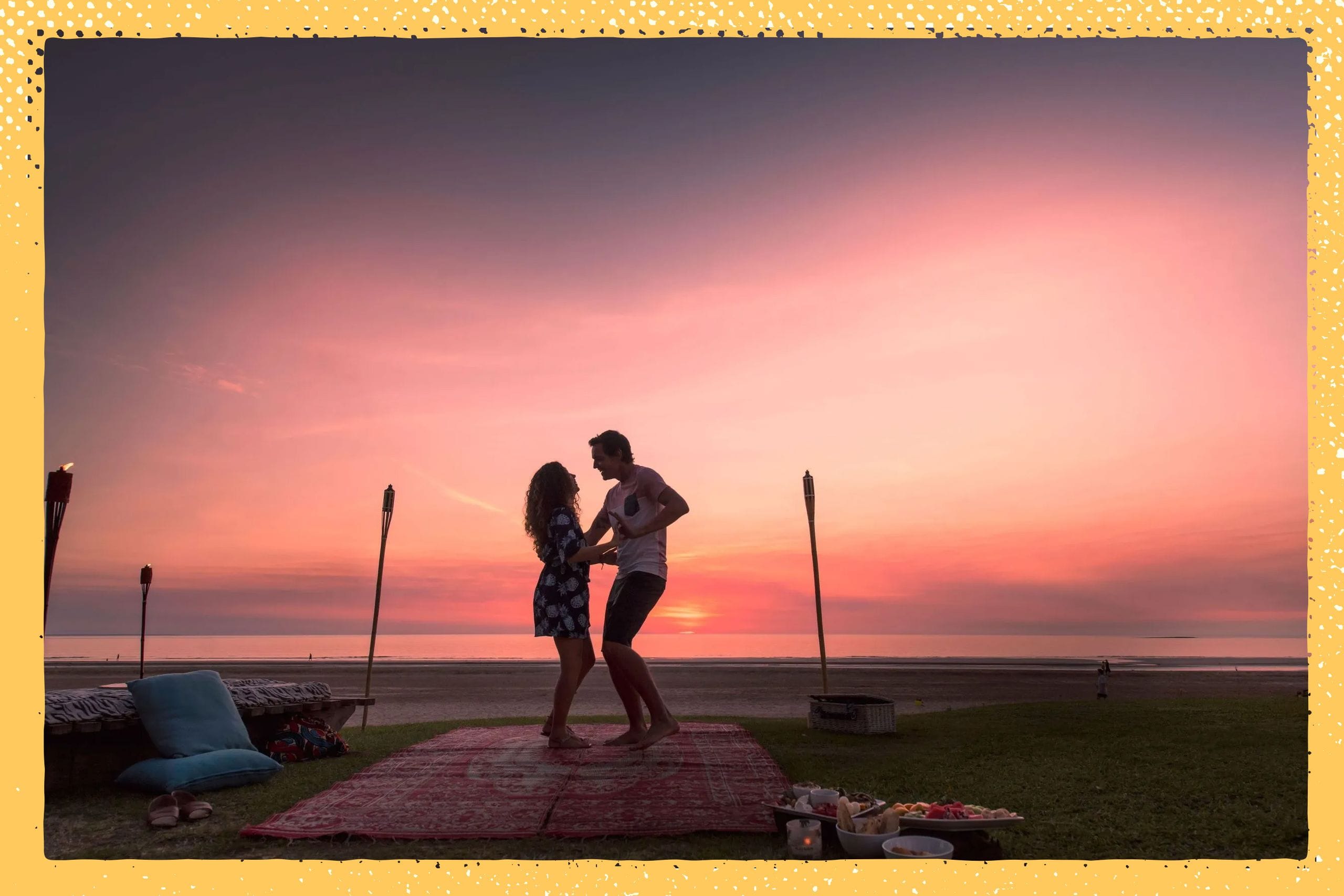 A couple dances on a beach at sunset, surrounded by torches and picnic items, with a vibrant pink and orange sky in the background.