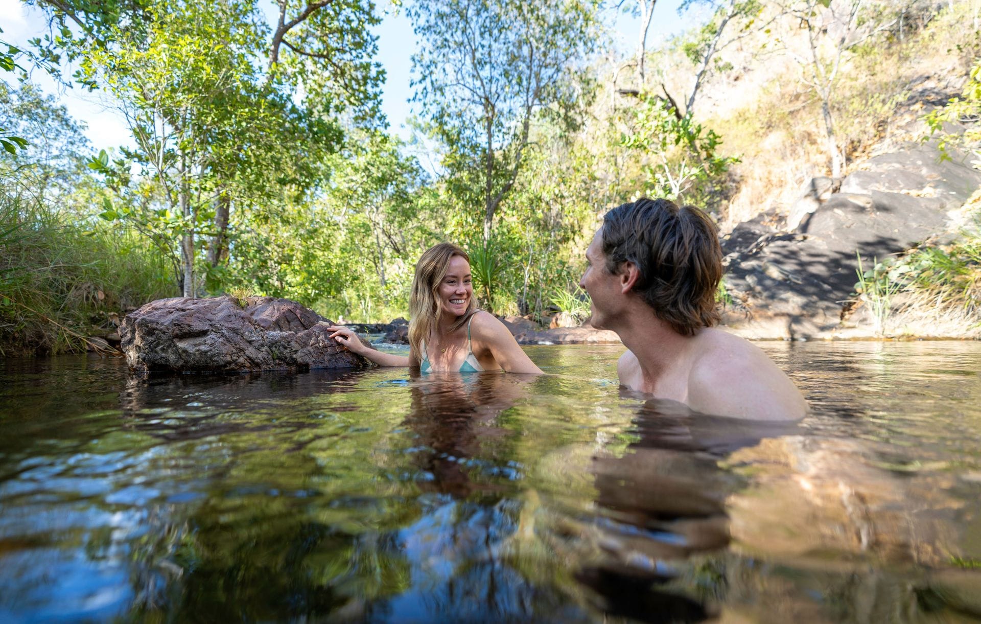 Two people swim and smile at each other in a clear, natural pool surrounded by lush greenery and rocky terrain.