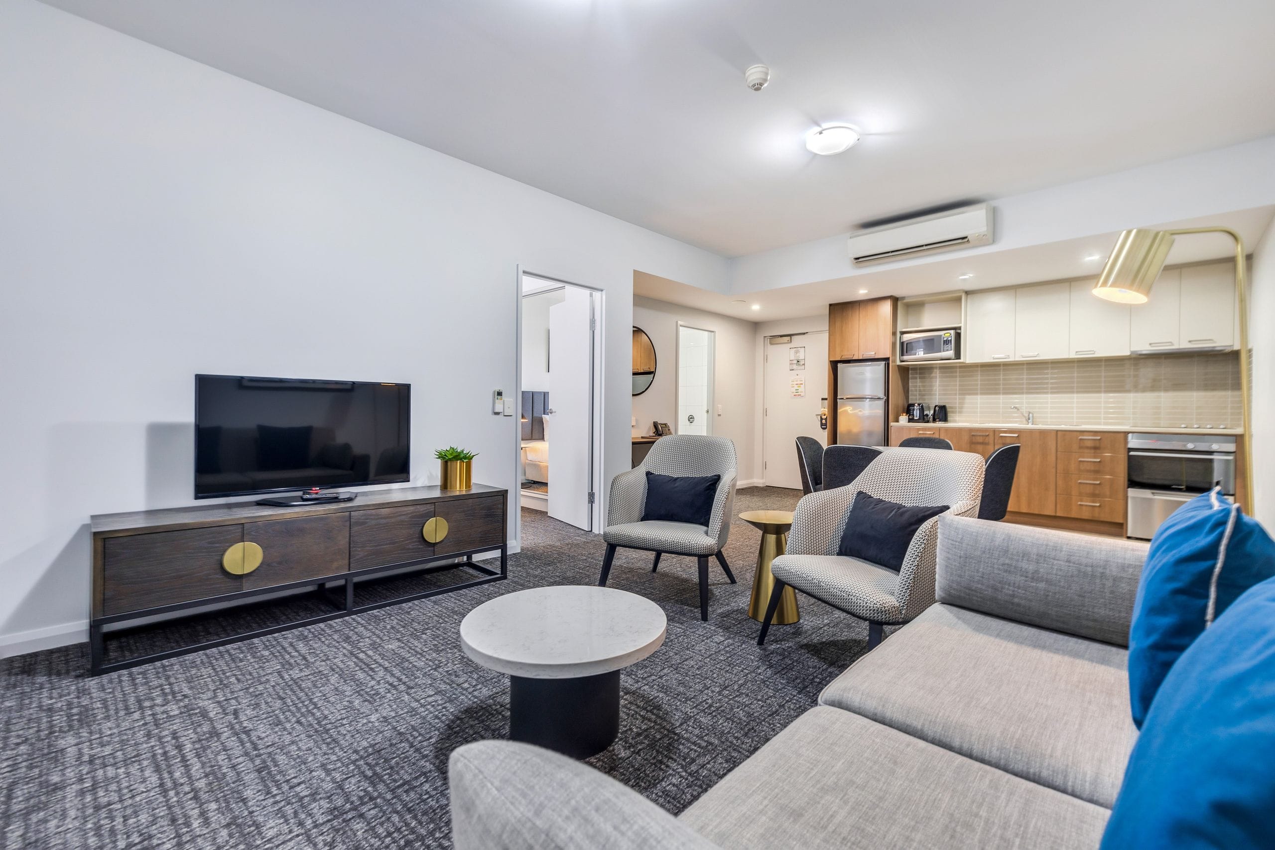 Modern living room with gray sofas, armchairs, flat-screen TV on a wooden console, round coffee table, and kitchenette in the background.