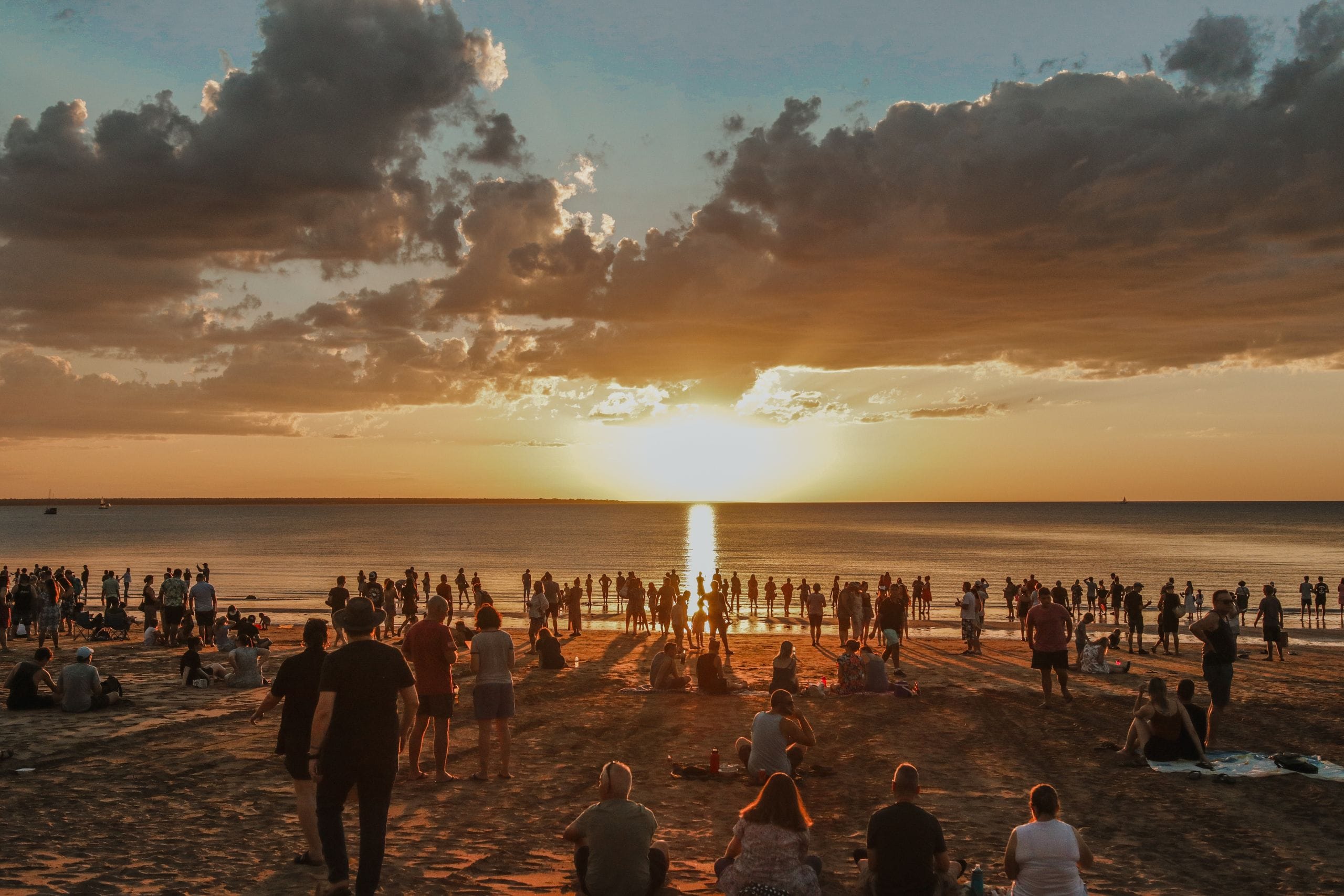 Crowd gathered on a beach watching a vibrant sunset over the ocean, with dramatic clouds and a golden reflection on the water.