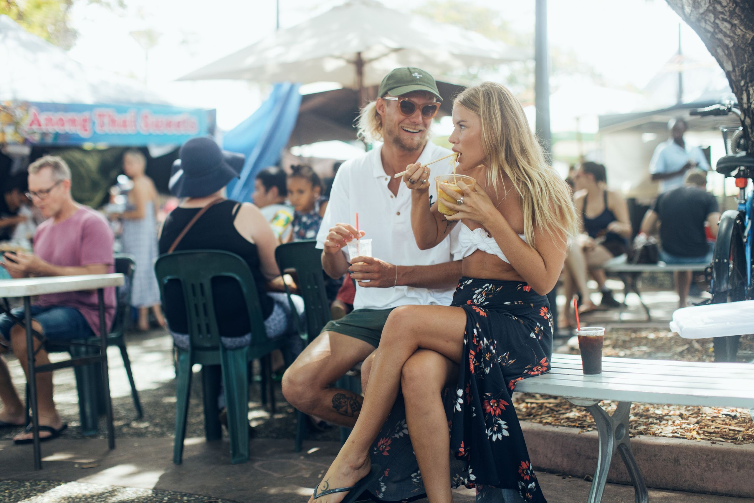 A couple sits on a bench enjoying drinks at an outdoor market, surrounded by people and umbrellas on a sunny day.